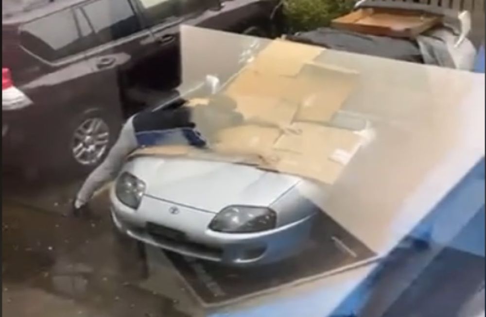 Al hombre se lo ve en el estacionamiento con su deportivo cubierto por cartones y abalanzándose arriba del vehículo tratando de cubrirlo de la lluvia con piedra. / Foto: Captura de video.