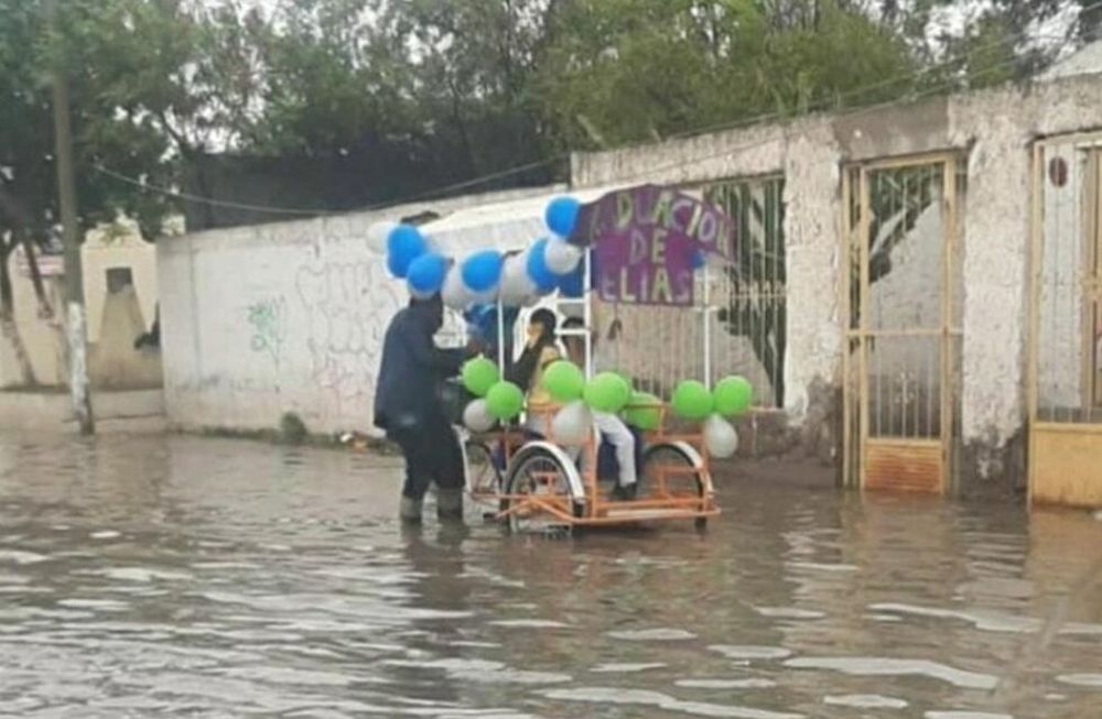 Un padre empuja un carro en medio del agua. Arriba del vehículo va su hijo, quien se recibió y ambos salieron a festejar por las calles.