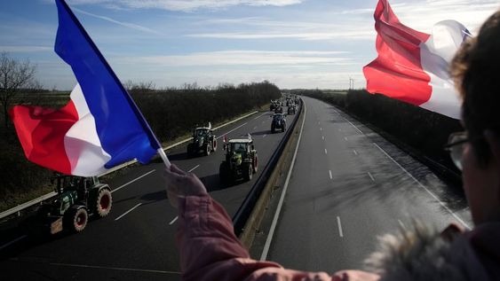 Una mujer con banderas aplaude al paso de campesinos en sus tractores que se dirigen a bloquear una autopista en Saint-Arnoult, al sur de París, viernes 26 de enero de 2024. Los agricultores bloquearon largos tramos de las principales autopistas de Francia el viernes, utilizando sus tractores para bloquear el tráfico y presionar al gobierno para que ceda a sus reclamos de facilitar y volver más rentable la producción de alimentos. (AP Foto/Christophe Ena)