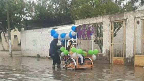 Un padre empuja un carro en medio del agua. Arriba del vehículo va su hijo, quien se recibió y ambos salieron a festejar por las calles.